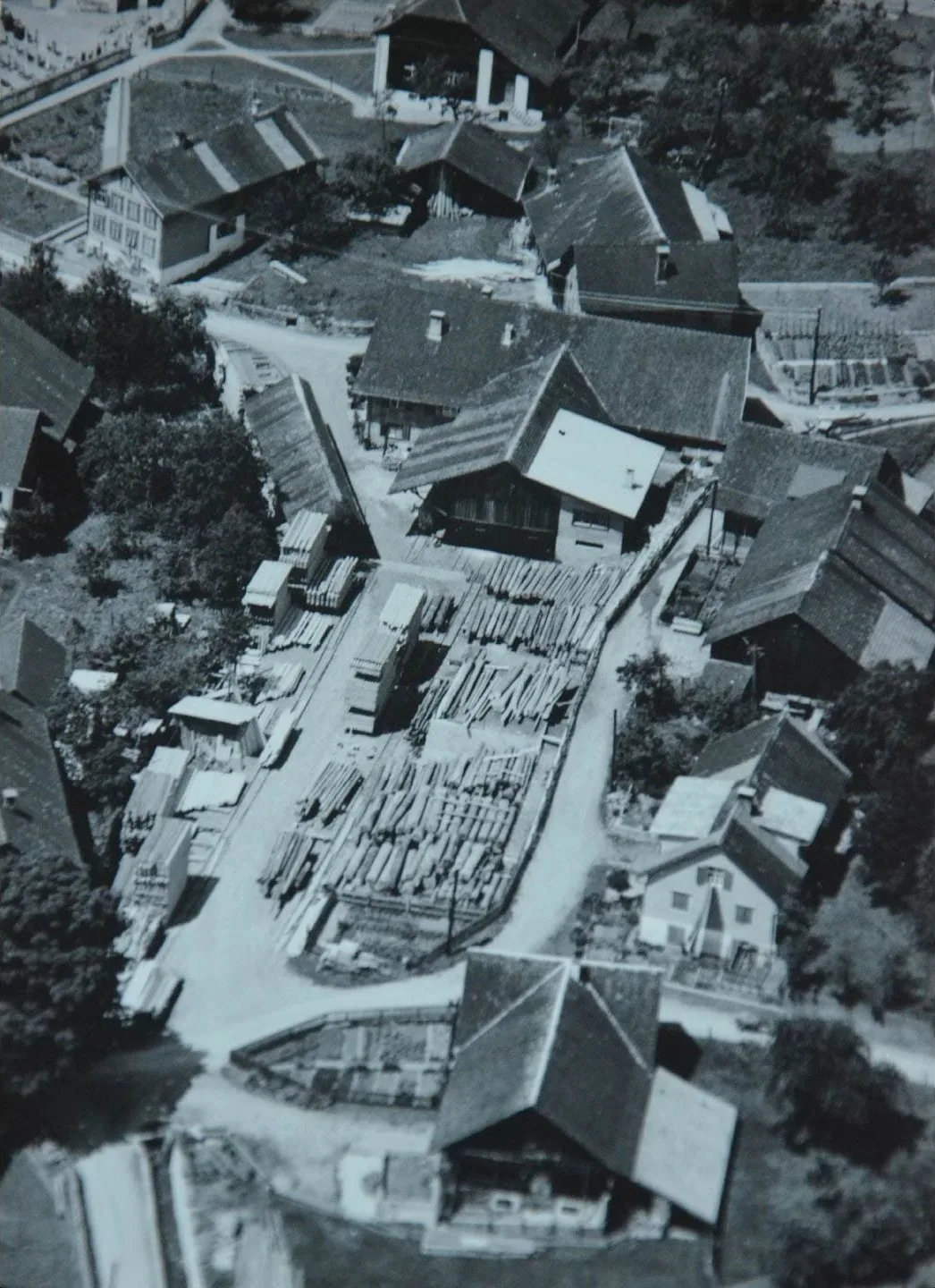 Schwarzweißes Luftbild von 1964: die alte Sägemühle mitten im Ortskern von Satteins, entlang der Gasse liegen Stämme und Schnittholzstapel zwischen den Wohnhäusern